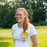 Glad kvinna med glasögon och rött hår ler i en vit t-shirt med ett trädmotiv. Hon verkar trivas med att jobba i naturen.
