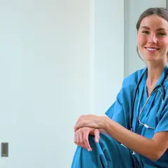 En leende sjuksköterska i blå uniform med stetoskop. Hon sitter avslappnat, en spegelbild av henne syns i bakgrunden. Ett inspirerande exempel på jobb inom vården.