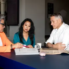 Tre personer diskuterar ekonom arbetsuppgifter vid ett bord med tidningar och dokument.
