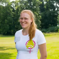 Glad kvinna med glasögon och rött hår ler i en vit t-shirt med ett trädmotiv. Hon verkar trivas med att jobba i naturen.