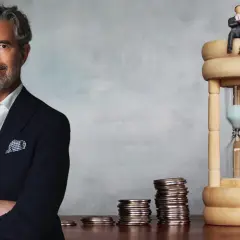 En man i kostym står bredvid en timglas och myntstaplar. Frågan är om man kan bli rik på aktier.