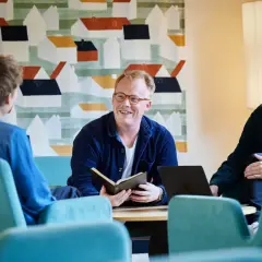 Tre män diskuterar vid ett bord, en med en öppen bok. Kanske en del av deras teologi utbildning.