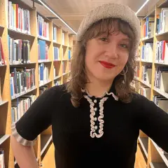En kvinna med mössa står bland bokhyllor. Hon ler och undrar hur blir man bibliotekarie?