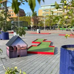 En minigolfbana i Helsingborg med en husmodell som visar stadens silhuett. Gröna träd och blå krukor ger liv åt platsen.