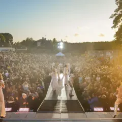 Stor publik njuter av Diggiloo Karlsborg i solnedgången. Artister på scenen framför en folkmassa.