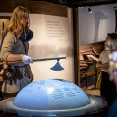 En guide i vikingakläder visar en yxa vid the viking museum. En jordglob med kartor och text projiceras.