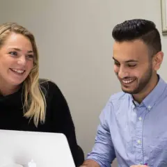 Två kollegor skrattar vid en laptop, kanske diskuterar de en ny grafisk design.