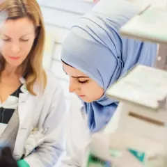 Två kvinnor i labbrockar, en med hijab, studerar något. De är på en arbetsplats som kan vara en del av en receptarie utbildning.