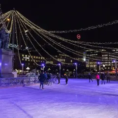 Människor åker skridskor på en upplyst isbana i Stockholm, omgivna av gnistrande ljusslingor och stadens silhuett.