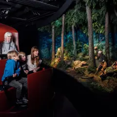Familj i vagn på vikingamuseum, tittar på en dramatisk scen med vikingar i skogen. Bra recensioner för detta museum!