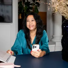 En leende kvinna med en telefon i handen, en bok och en rosa högtalare på bordet. Hon är en framgångsrik ekonomi konsult.