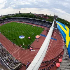 Stor svensk flagga vajar över en fullsatt arena i Stockholm. En friidrottstävling pågår på den röda löparbanan.