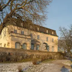 Prins Eugens Waldemarsudde i vinterskrud, med en stor ek och en staty framför den gula byggnaden.
