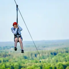 Glad flicka i hjälm susar nerför lina över skog i Skåne. En perfekt barnaktivitet för äventyrliga barn.