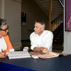 Två personer diskuterar vid ett bord, en kvinna med orange kavaj antecknar, en man pekar på en tidning. Kanske ett möte om administrativa jobb.
