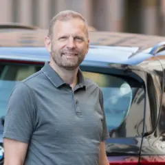 En glad man står bredvid en bil. Kanske funderar han på att leasa en begagnad bil för en bekväm resa.