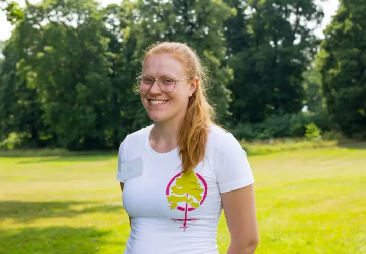 Glad kvinna med glasögon och rött hår ler i en vit t-shirt med ett trädmotiv. Hon verkar trivas med att jobba i naturen.