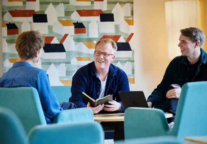 Tre män diskuterar vid ett bord, en med en öppen bok. Kanske en del av deras teologi utbildning.