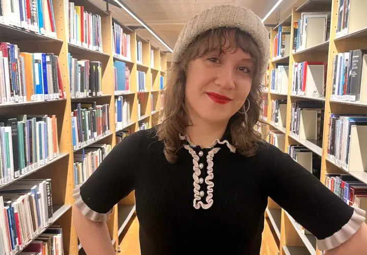 En kvinna med mössa står bland bokhyllor. Hon ler och undrar hur blir man bibliotekarie?