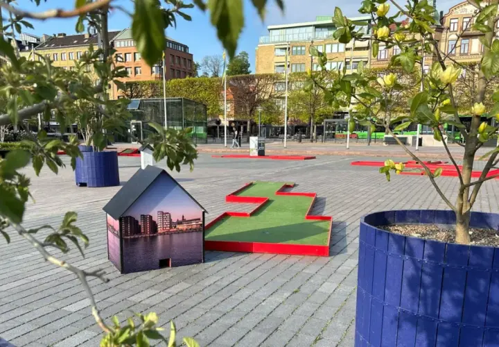 En minigolfbana i Helsingborg med en husmodell som visar stadens silhuett. Gröna träd och blå krukor ger liv åt platsen.