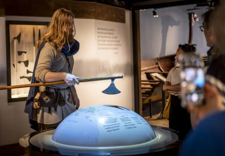 En guide i vikingakläder visar en yxa vid the viking museum. En jordglob med kartor och text projiceras.
