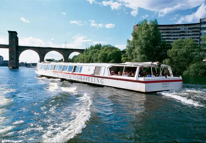 Sightseeingbåt passerar under en gammal tegelbro i Stockholm. Vattnet skummar bakom båten.