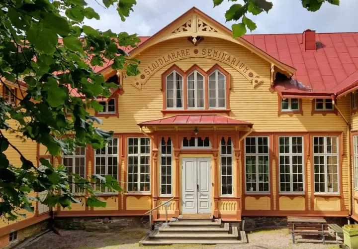 Gult trähus med röda detaljer, "Slöjdlärare Seminarium" text. Perfekt för en byggnadsvård kurs distans.