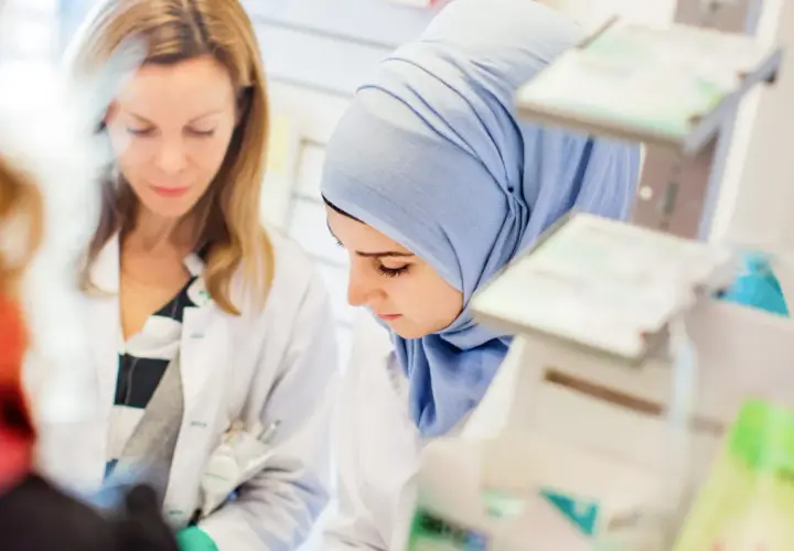 Två kvinnor i labbrockar, en med hijab, studerar något. De är på en arbetsplats som kan vara en del av en receptarie utbildning.