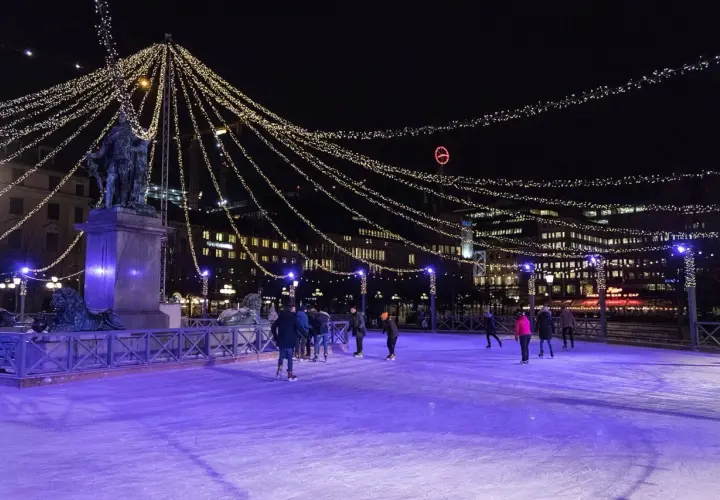 Människor åker skridskor på en upplyst isbana i Stockholm, omgivna av gnistrande ljusslingor och stadens silhuett.