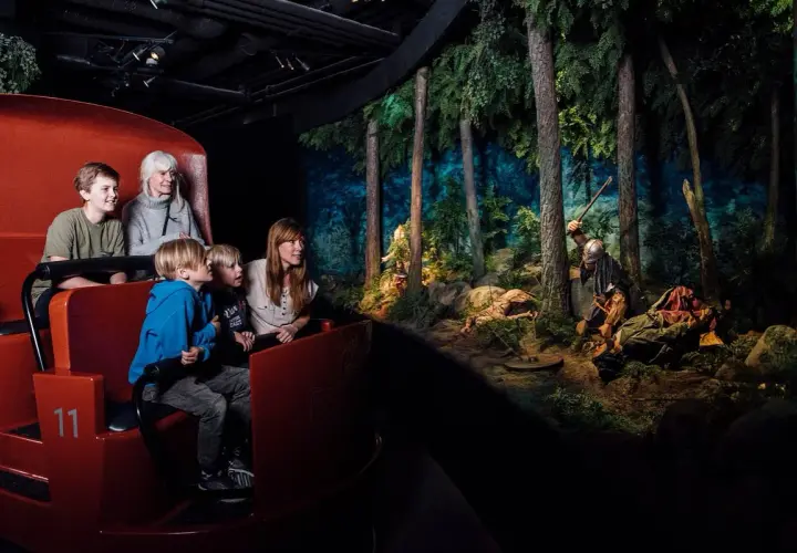Familj i vagn på vikingamuseum, tittar på en dramatisk scen med vikingar i skogen. Bra recensioner för detta museum!