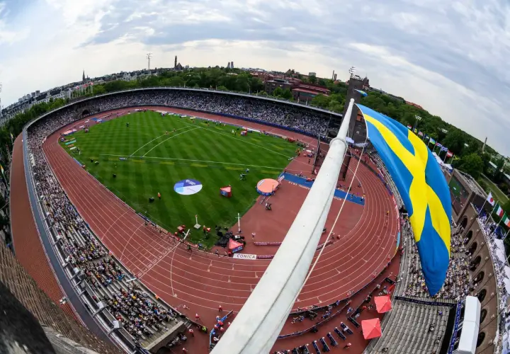 Stor svensk flagga vajar över en fullsatt arena i Stockholm. En friidrottstävling pågår på den röda löparbanan.