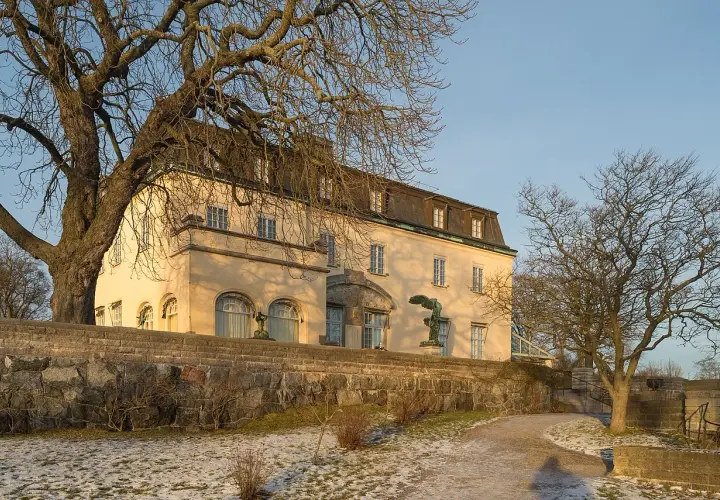 Prins Eugens Waldemarsudde i vinterskrud, med en stor ek och en staty framför den gula byggnaden.