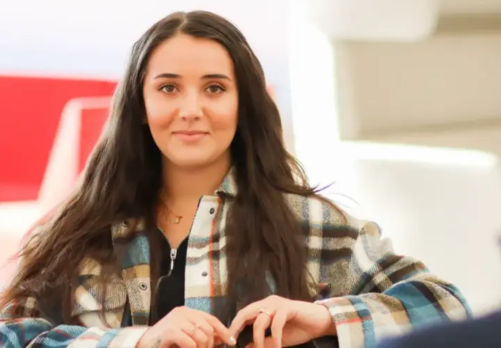Ung kvinna med mörkt hår, klädd i rutig skjorta, ler mot kameran. Hon kanske studerar socialpedagog utbildning distans.