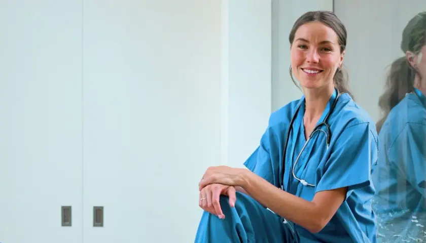 En leende sjuksköterska i blå uniform med stetoskop. Hon sitter avslappnat, en spegelbild av henne syns i bakgrunden. Ett inspirerande exempel på jobb inom vården.