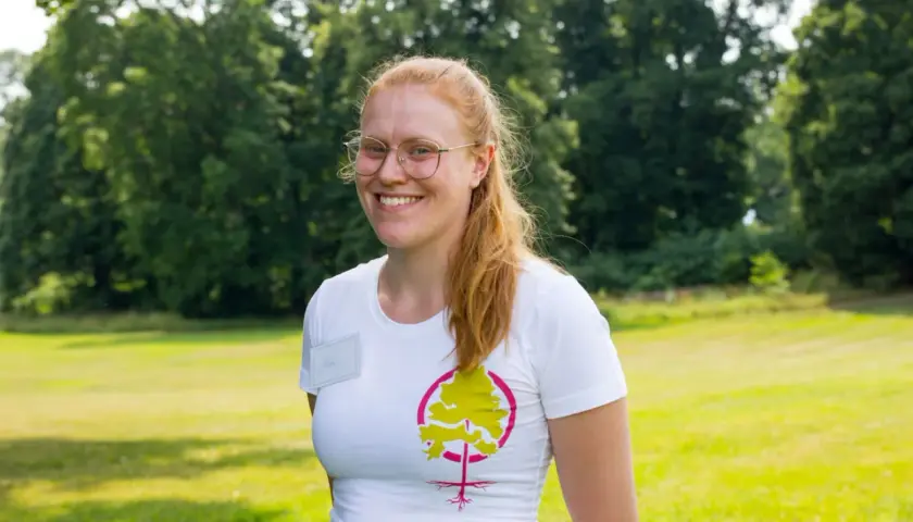 Glad kvinna med glasögon och rött hår ler i en vit t-shirt med ett trädmotiv. Hon verkar trivas med att jobba i naturen.