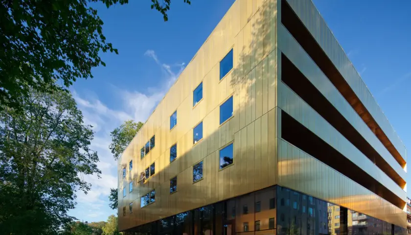 Sven-Harrys konstmuseum glänser i guld under en klarblå himmel. Gröna träd ramar in den moderna byggnaden.