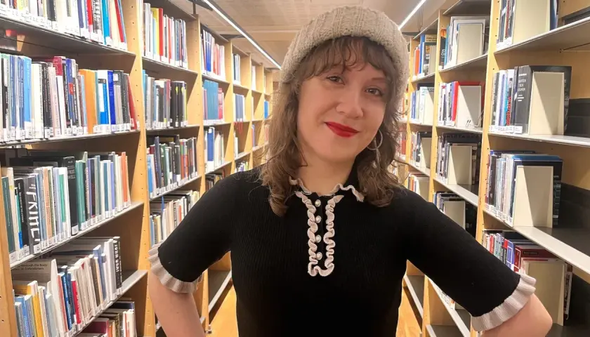 En kvinna med mössa står bland bokhyllor. Hon ler och undrar hur blir man bibliotekarie?