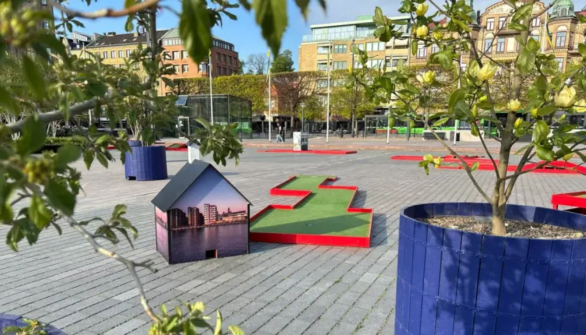 En minigolfbana i Helsingborg med en husmodell som visar stadens silhuett. Gröna träd och blå krukor ger liv åt platsen.