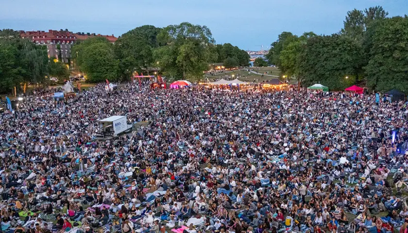 Stor publik samlad för utomhusbio i Stockholm. Tusentals människor sitter på filtar i en park och tittar mot en scen.