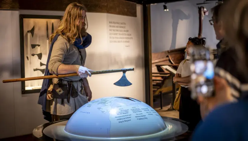 En guide i vikingakläder visar en yxa vid the viking museum. En jordglob med kartor och text projiceras.