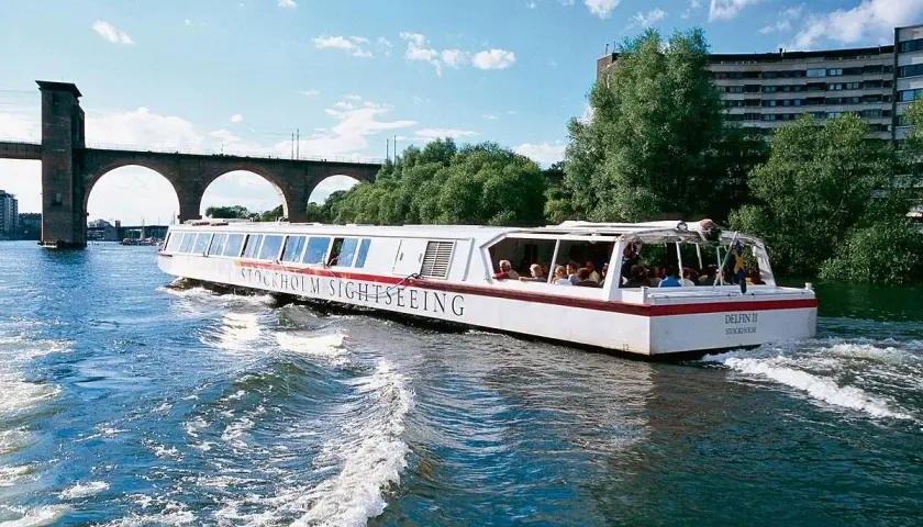Sightseeingbåt passerar under en gammal tegelbro i Stockholm. Vattnet skummar bakom båten.