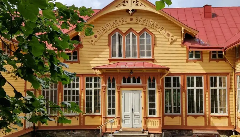 Gult trähus med röda detaljer, "Slöjdlärare Seminarium" text. Perfekt för en byggnadsvård kurs distans.