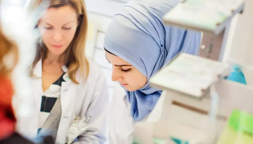 Två kvinnor i labbrockar, en med hijab, studerar något. De är på en arbetsplats som kan vara en del av en receptarie utbildning.