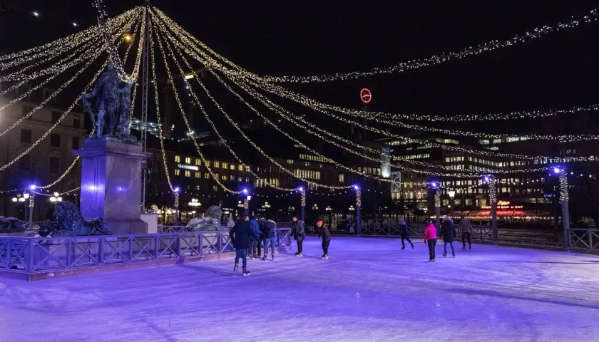 Människor åker skridskor på en upplyst isbana i Stockholm, omgivna av gnistrande ljusslingor och stadens silhuett.