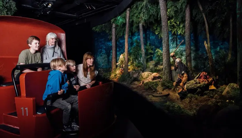 Familj i vagn på vikingamuseum, tittar på en dramatisk scen med vikingar i skogen. Bra recensioner för detta museum!