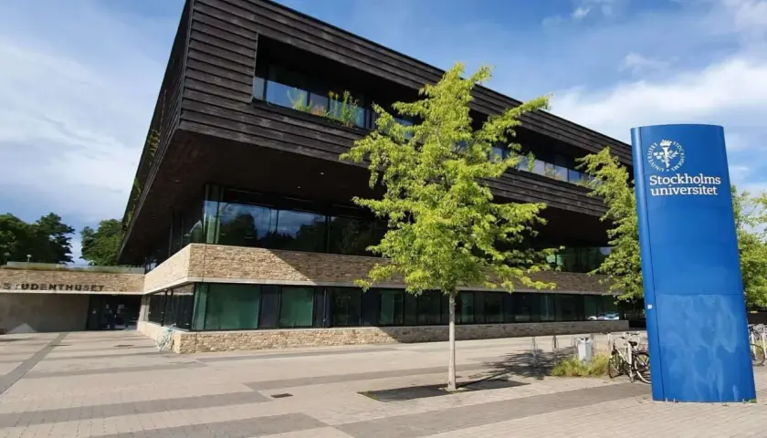 Modern byggnad med stenfasad och mörka träpaneler. En blå skylt med "Stockholms universitet" står bredvid. En del av universitet i Stockholm lista.