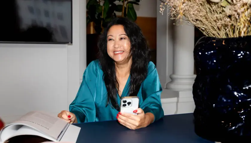 En leende kvinna med en telefon i handen, en bok och en rosa högtalare på bordet. Hon är en framgångsrik ekonomi konsult.