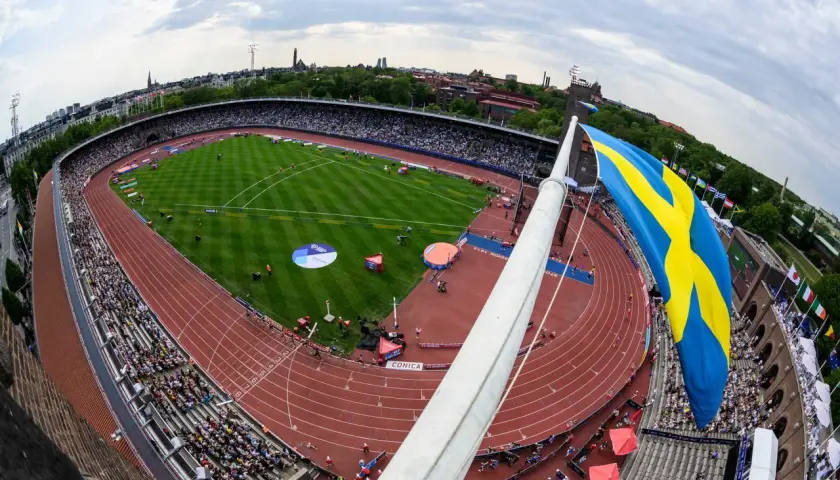 Stor svensk flagga vajar över en fullsatt arena i Stockholm. En friidrottstävling pågår på den röda löparbanan.