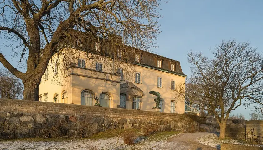 Prins Eugens Waldemarsudde i vinterskrud, med en stor ek och en staty framför den gula byggnaden.