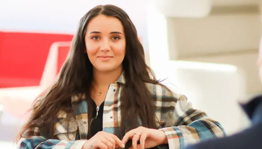 Ung kvinna med mörkt hår, klädd i rutig skjorta, ler mot kameran. Hon kanske studerar socialpedagog utbildning distans.